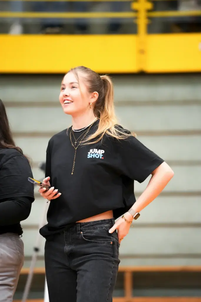Lachende blonde Frau in schwarzem Shirt „JUMP SHOT.“ hält ein Gerät. Optimal für Social Media oder Medienagentur.