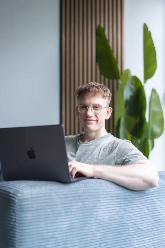 Junger Mann mit Brille sitzt an einem Laptop vor einem hellblauen Sofa; grünes Pflanzenblatt im Hintergrund.