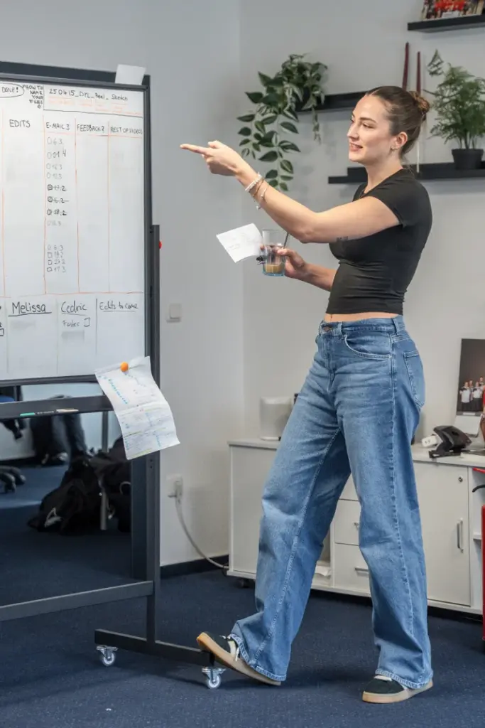 Frau in Büro diskutiert Content-Planung an einem Whiteboard. Sie zeigt auf eine Tabelle mit Stichpunkten zu Social Media und Feedback.
