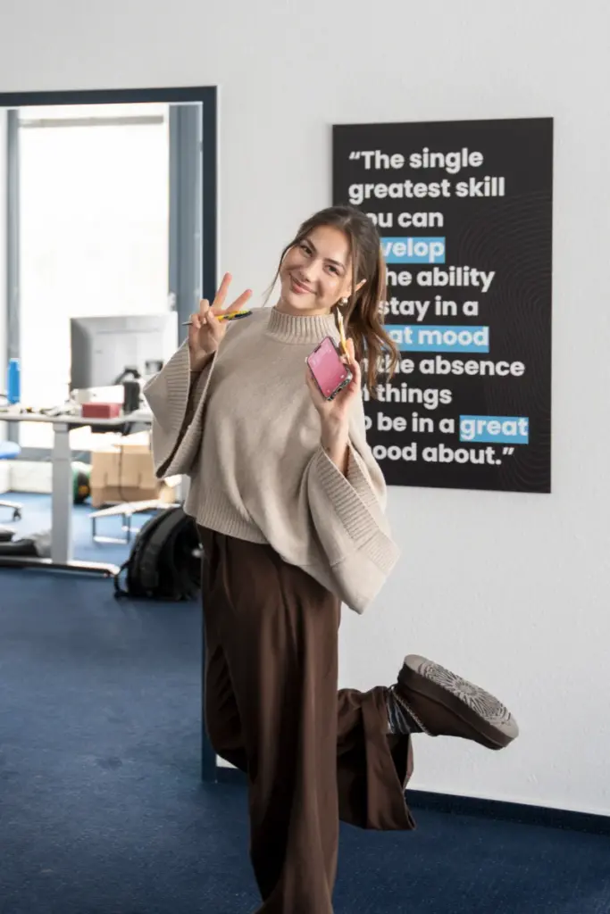 Lächelnde Frau mit V-Zeichen in einem modernen Büro. Poster und Dekoration deuten auf eine Medienagentur in Paderborn hin.