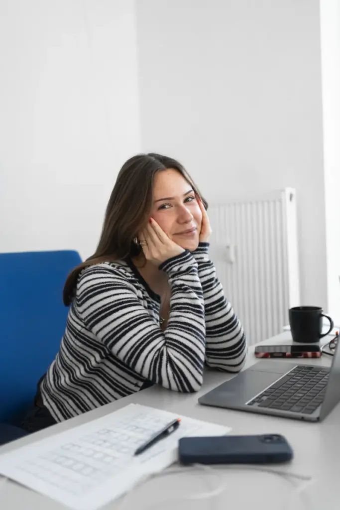 Frau an Schreibtisch mit Notebook, Smartphone und Tasse vor weißer Wand in hellem Büro.