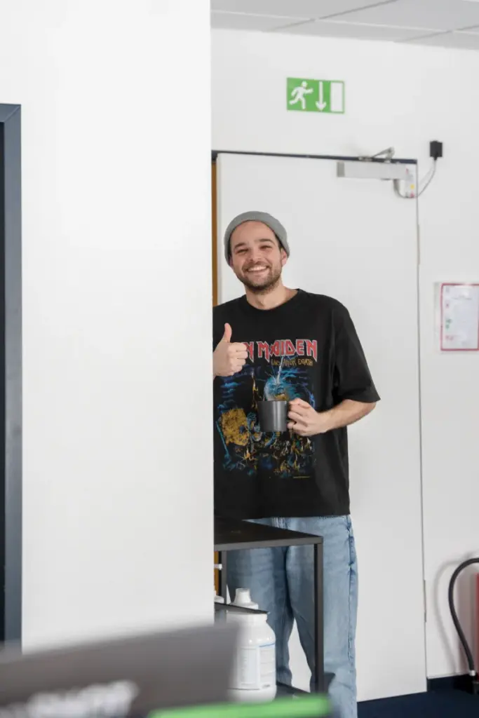 Mann in einem modernen Büroflur einer Medienagentur. Er lächelt, macht Daumen hoch und trägt dunkles T-Shirt.